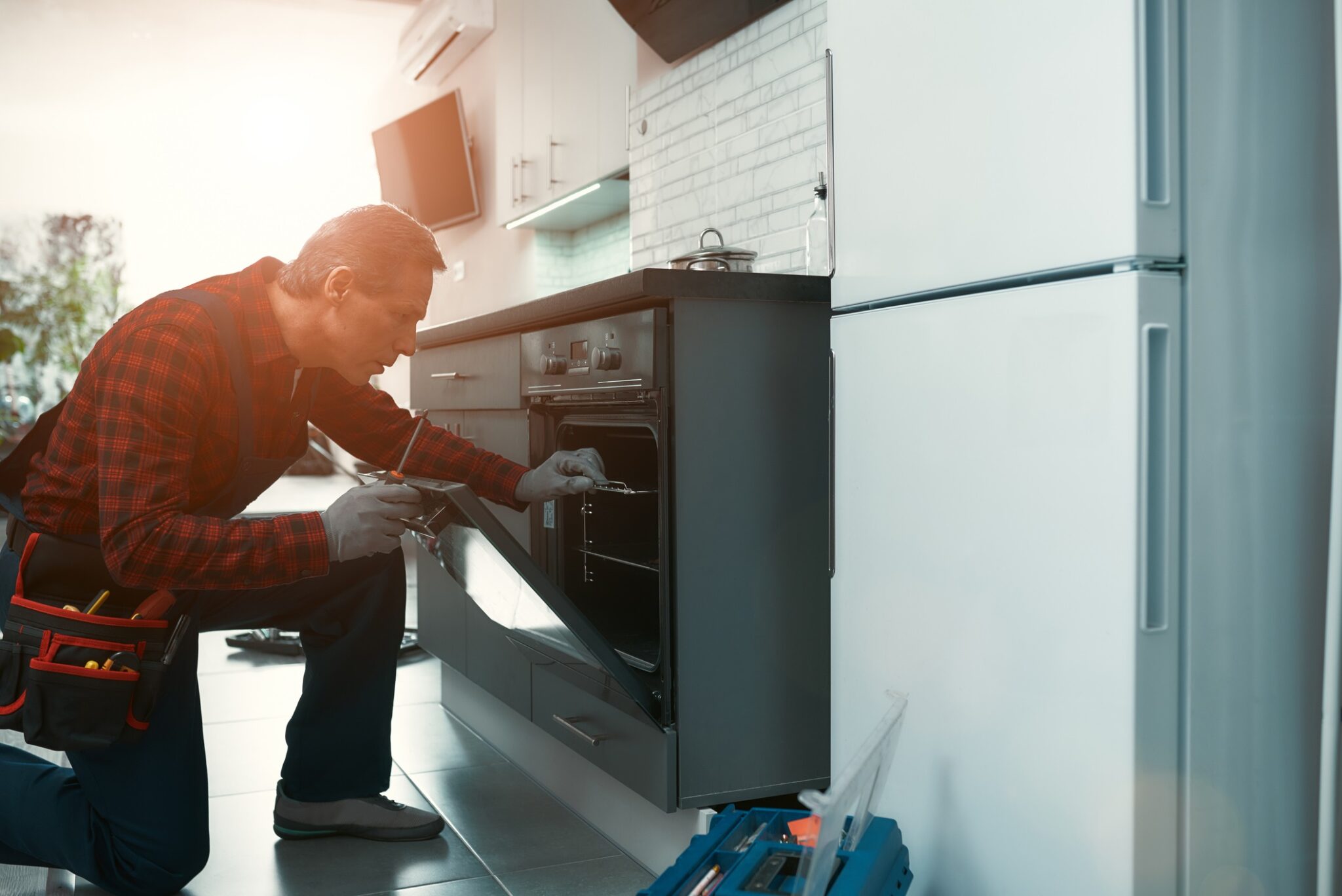 A man wearing a tool kit works on repairing an oven for home services company blog.