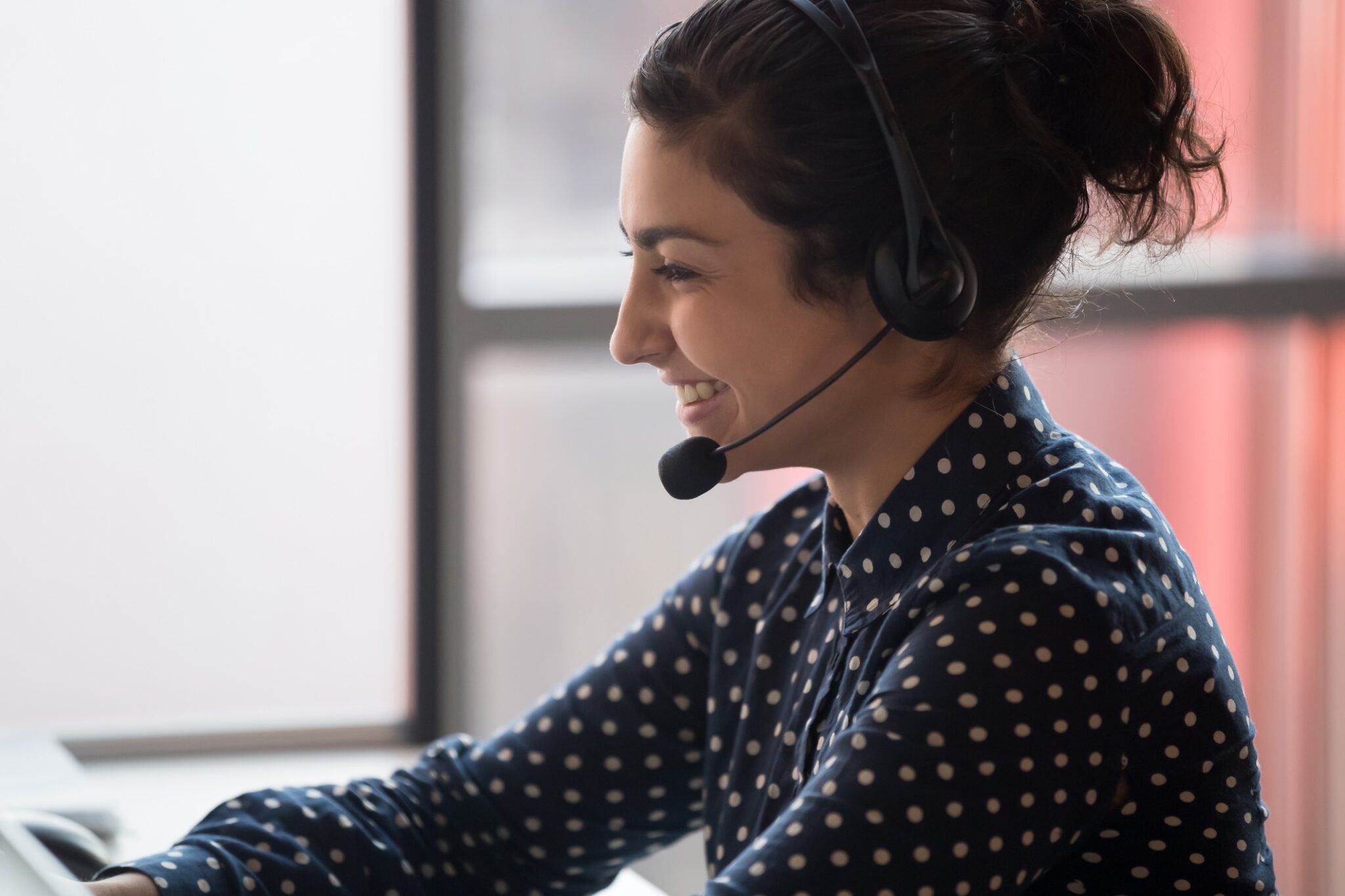 A young female call center agent wearing a headset smiles as she consults a client online for specialization in business blog.