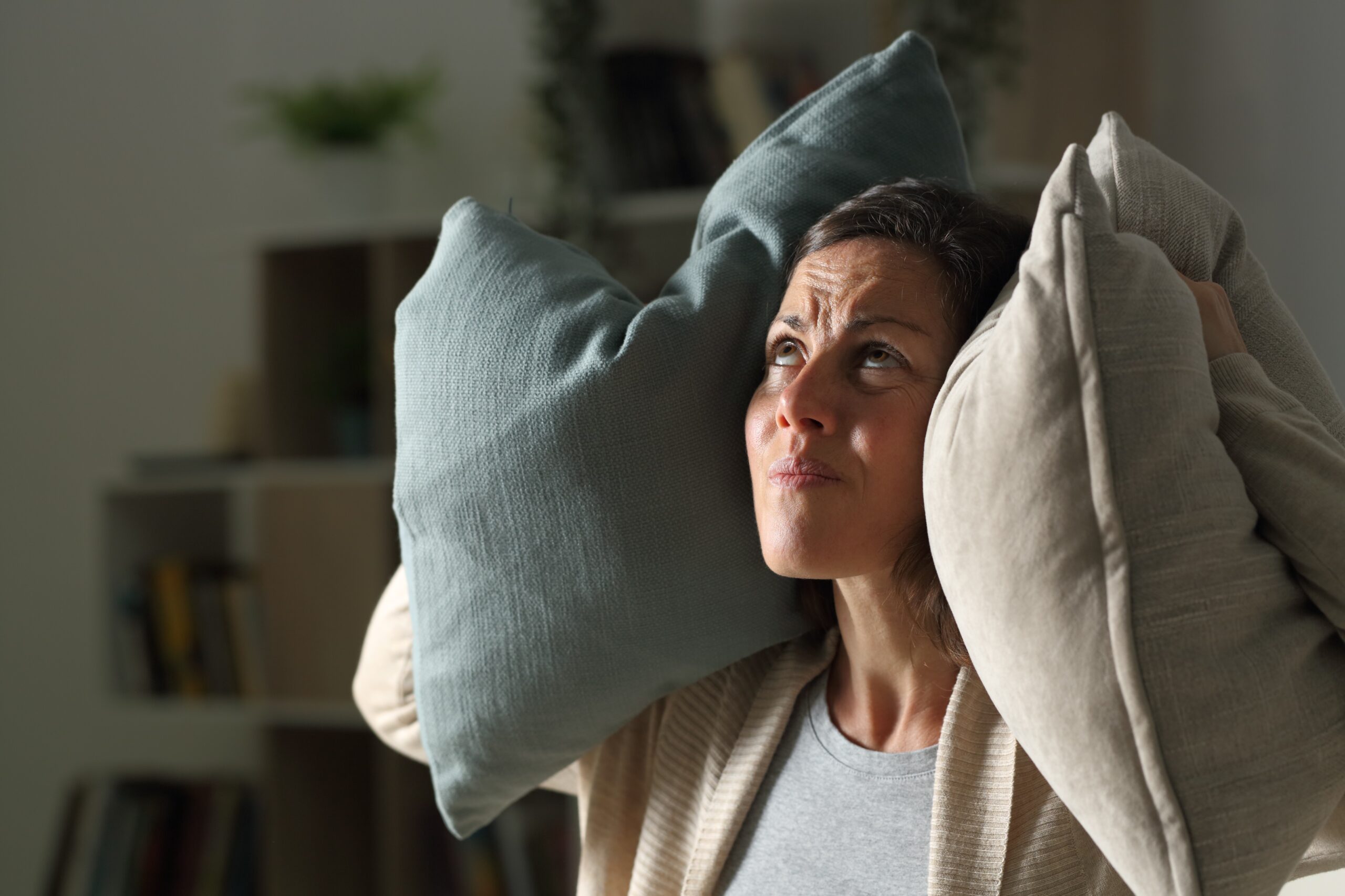 An annoyed woman covers her ears with pillows and stares at toward the apartment above with a frustrated expression for noise complaints from residents blog.