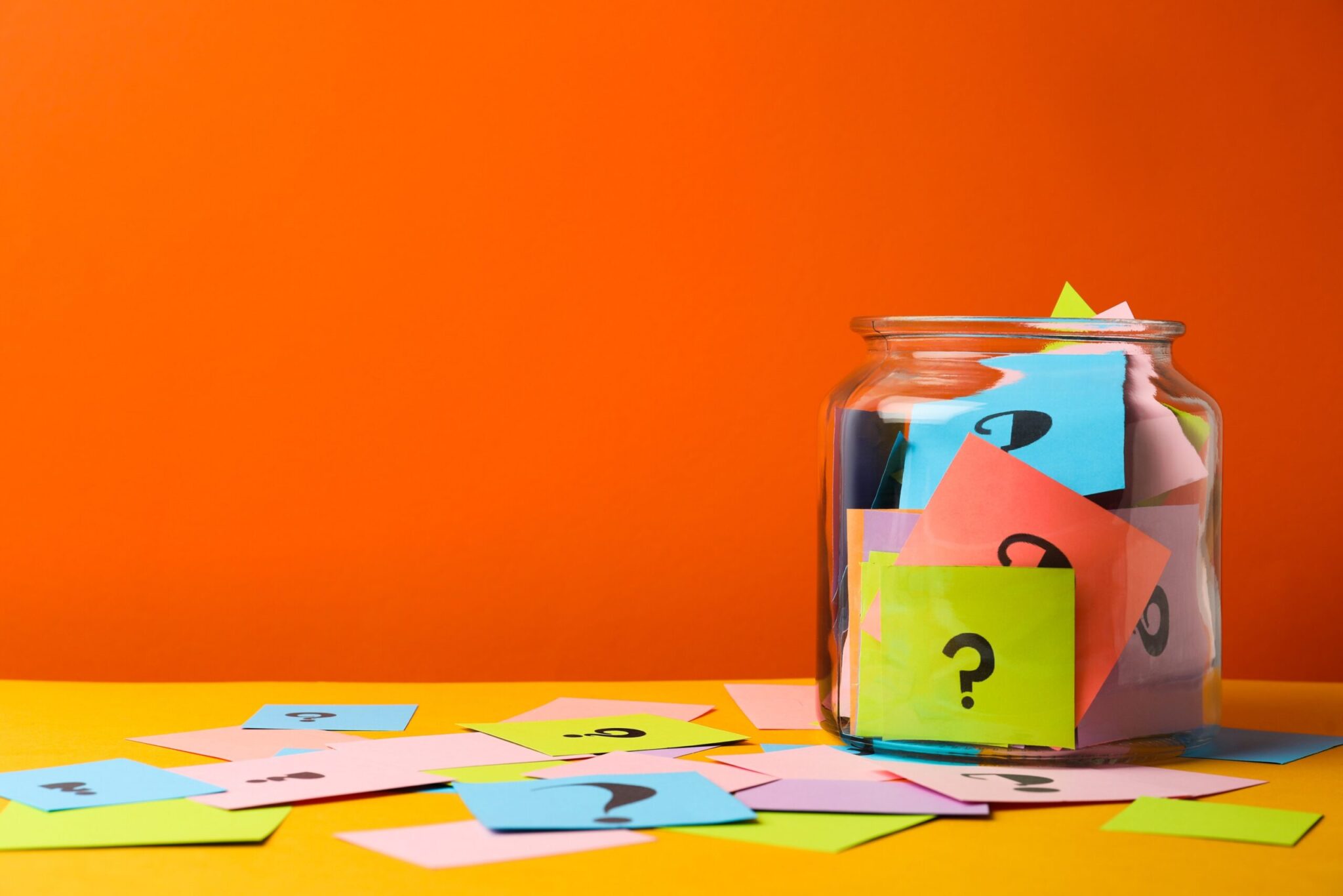 A glass jar filled with multicolor cards with question marks on them is seated on a yellow table, littered with similar cards, against an orange background for which questions do customers want answered blog.