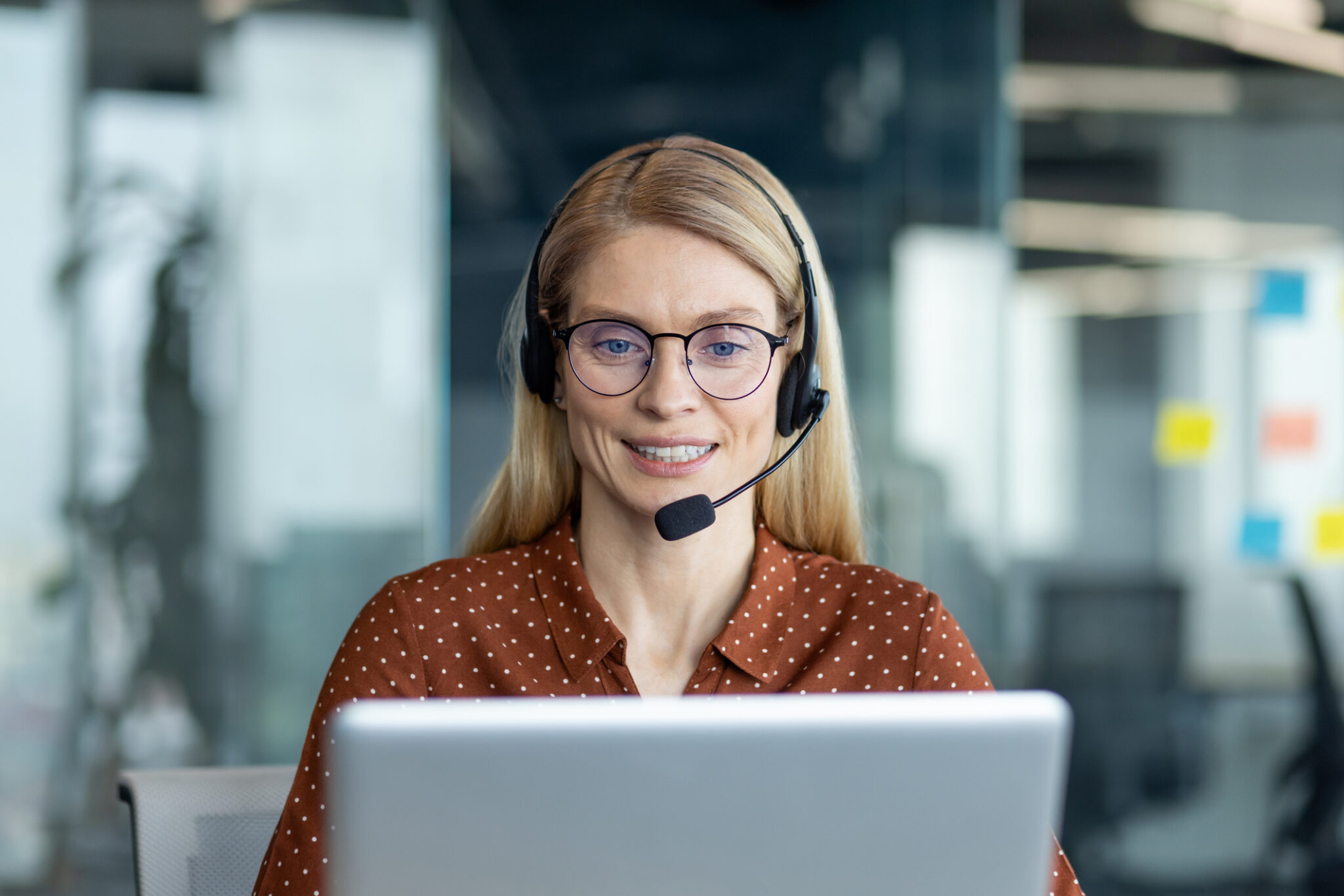 A friendly answering service agent smiles while wearing a headset and working on a laptop for franchise-answering-service blog.