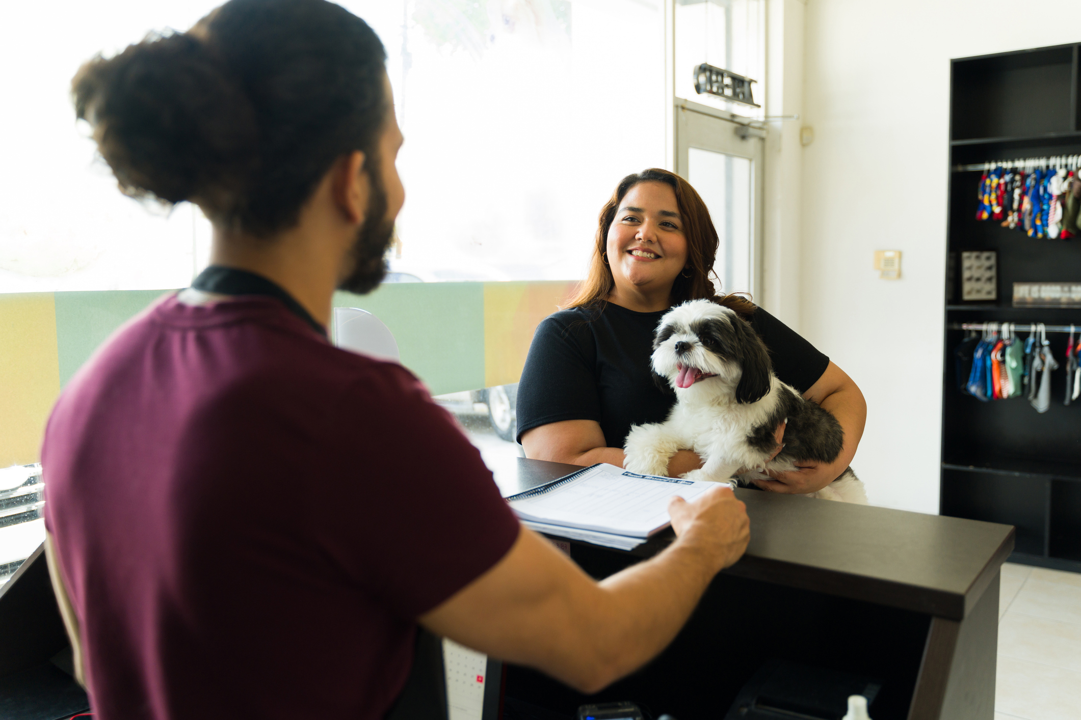 Smiling pet parent holding a shih tzu at a pet care front desk while a staff member reviews check-in paperwork for peak-season-staffing-for-pet-care blog.